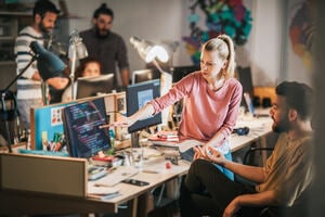 Group of computer programmers working in the office. Focus is on blond woman showing something to her colleague on PC relating to code.